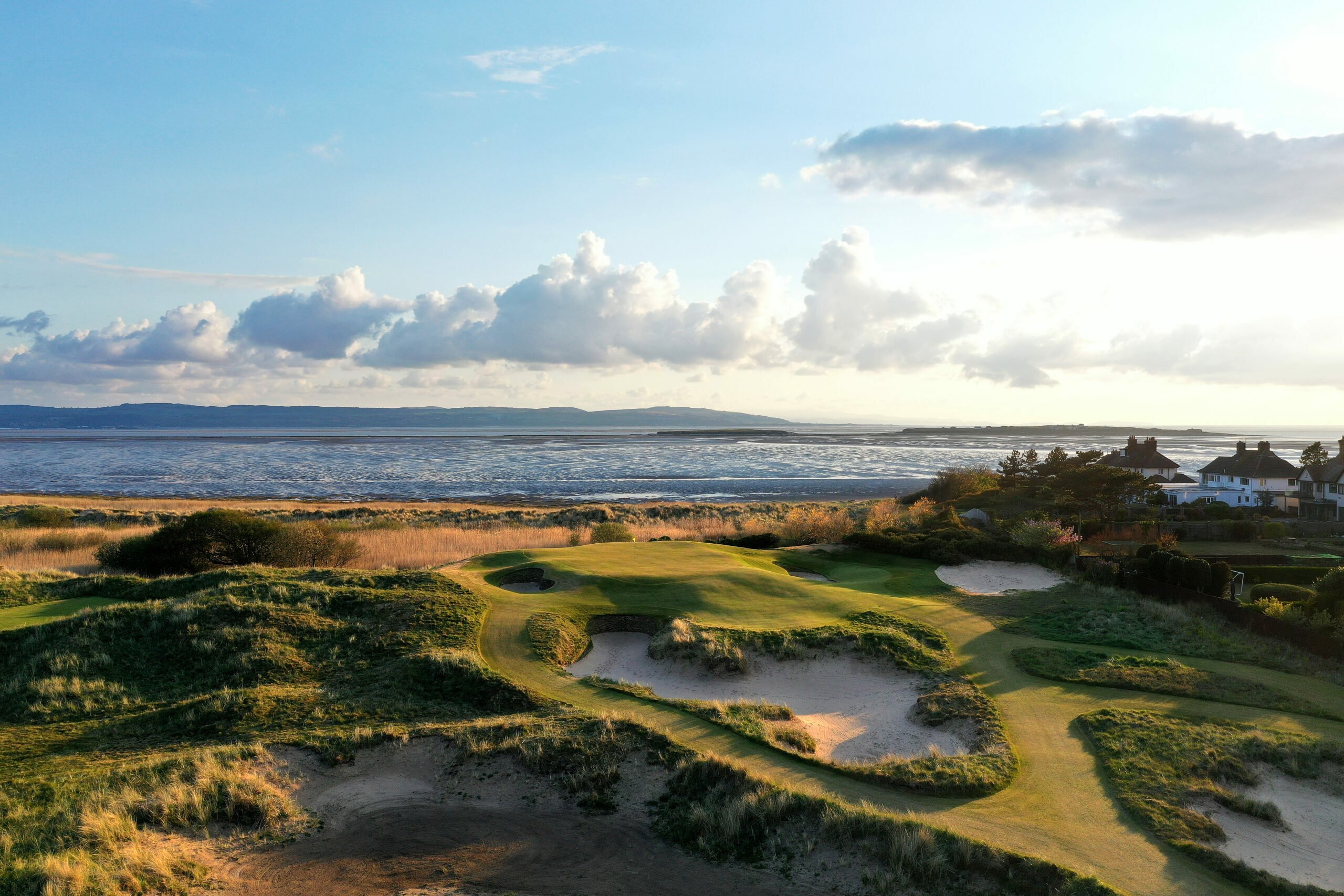 A scenic coastal golf course with rolling grassy dunes and sandy bunkers in the foreground, overlooking a calm sea under a partly cloudy sky. Houses are visible to the right, and distant hills stretch across the horizon.