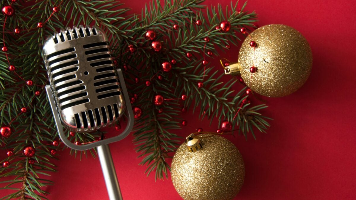 Vintage silver microphone placed on a red background with green pine branches, red berries, and two glittery gold Christmas ornaments.