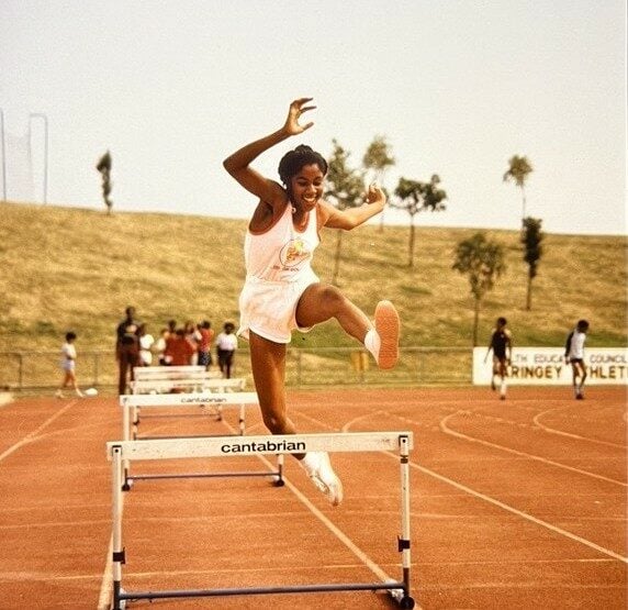 An athlete in mid-air clearing a hurdle on a red running track during a race. The athlete is wearing a white athletic uniform with a logo on the chest and white shoes. The hurdle is labeled “cantabrian.” In the background, there are several people standing near the track and grassy slopes with scattered trees under a clear sky.