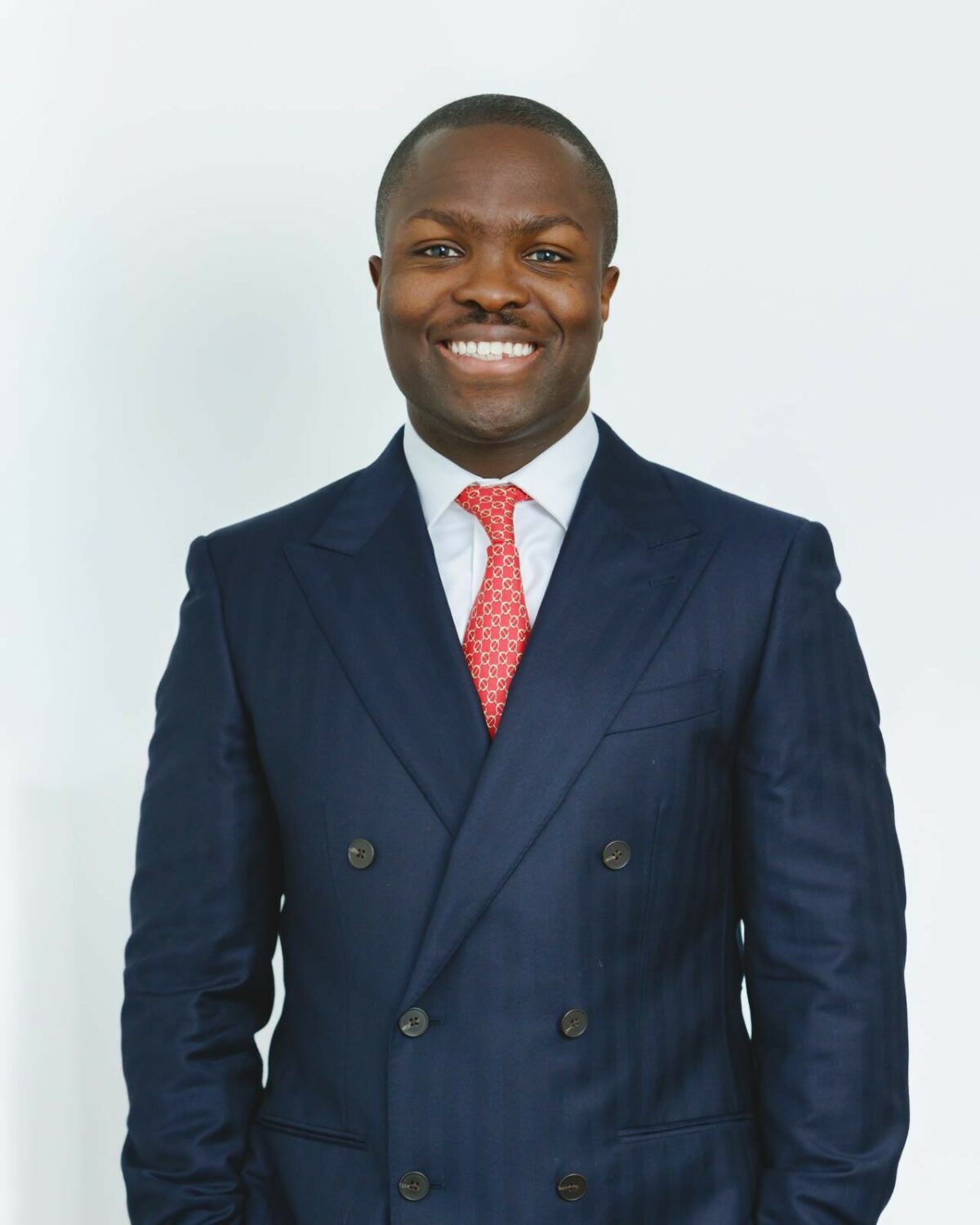 Ofei Kwafo-Akoto wearing a dark navy double-breasted suit with peak lapels, paired with a white dress shirt and a red patterned tie, standing against a plain white background.
