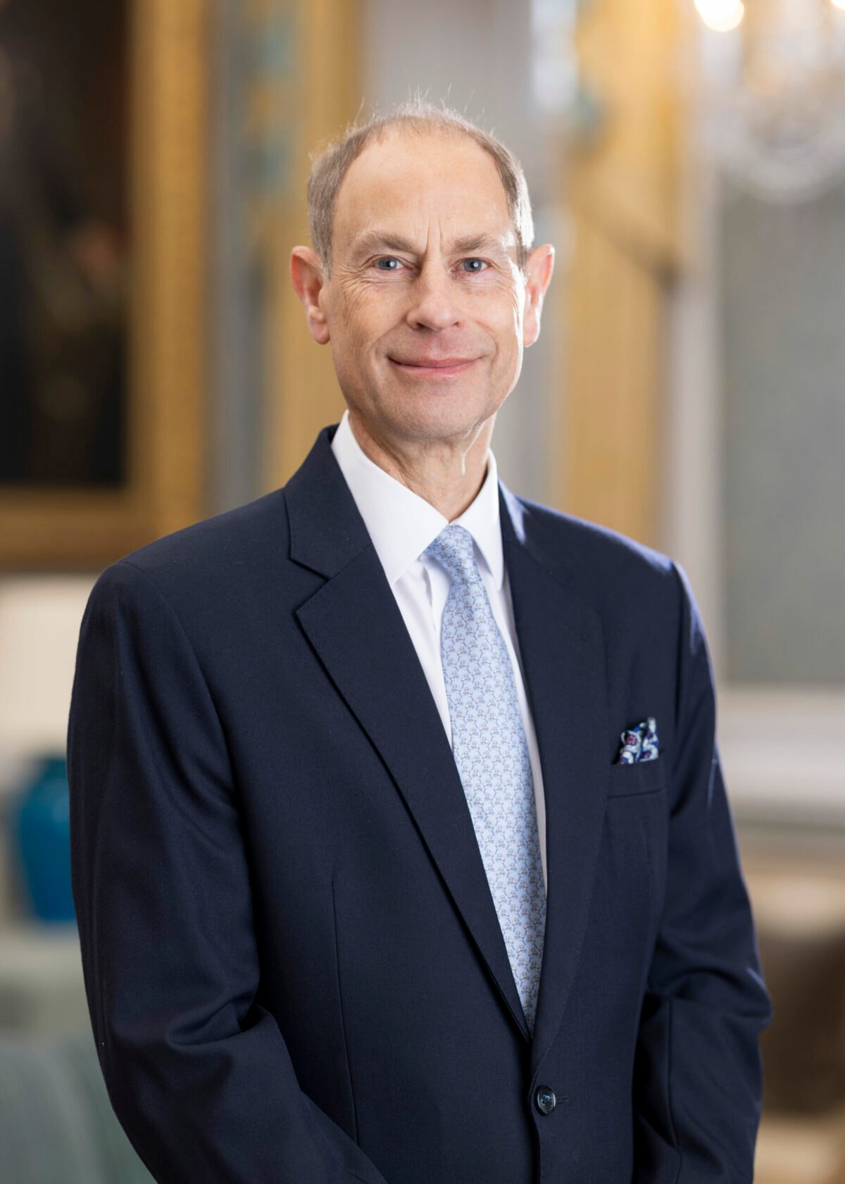 HRH The Duke of Edinburgh wearing a dark navy suit with a light blue patterned tie and a white dress shirt. A pocket square is visible in the breast pocket. The background shows an elegant interior with ornate details, including a chandelier and framed artwork.