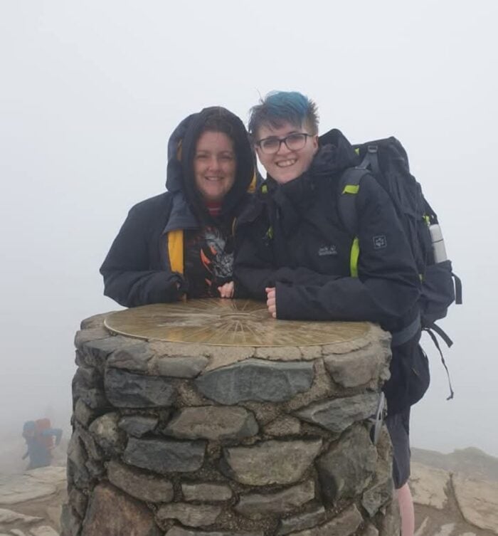 Michelle and Jay standing together at a stone summit marker on a mountain, wearing dark outdoor jackets and backpacks. The scene is surrounded by thick fog, creating a misty and atmospheric backdrop.