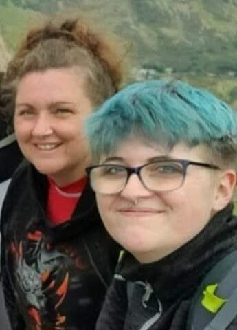 Michelle and Jay standing outdoors with a mountainous landscape in the background. The hills are green with rocky patches, suggesting a rugged terrain. Michelle is wearing a black hoodie with a colorful dragon design and a red shirt underneath. Jay has bright teal-colored hair and is dressed in a dark jacket with a backpack featuring a neon yellow strap. The photo appears to be taken during a hike or outdoor adventure.