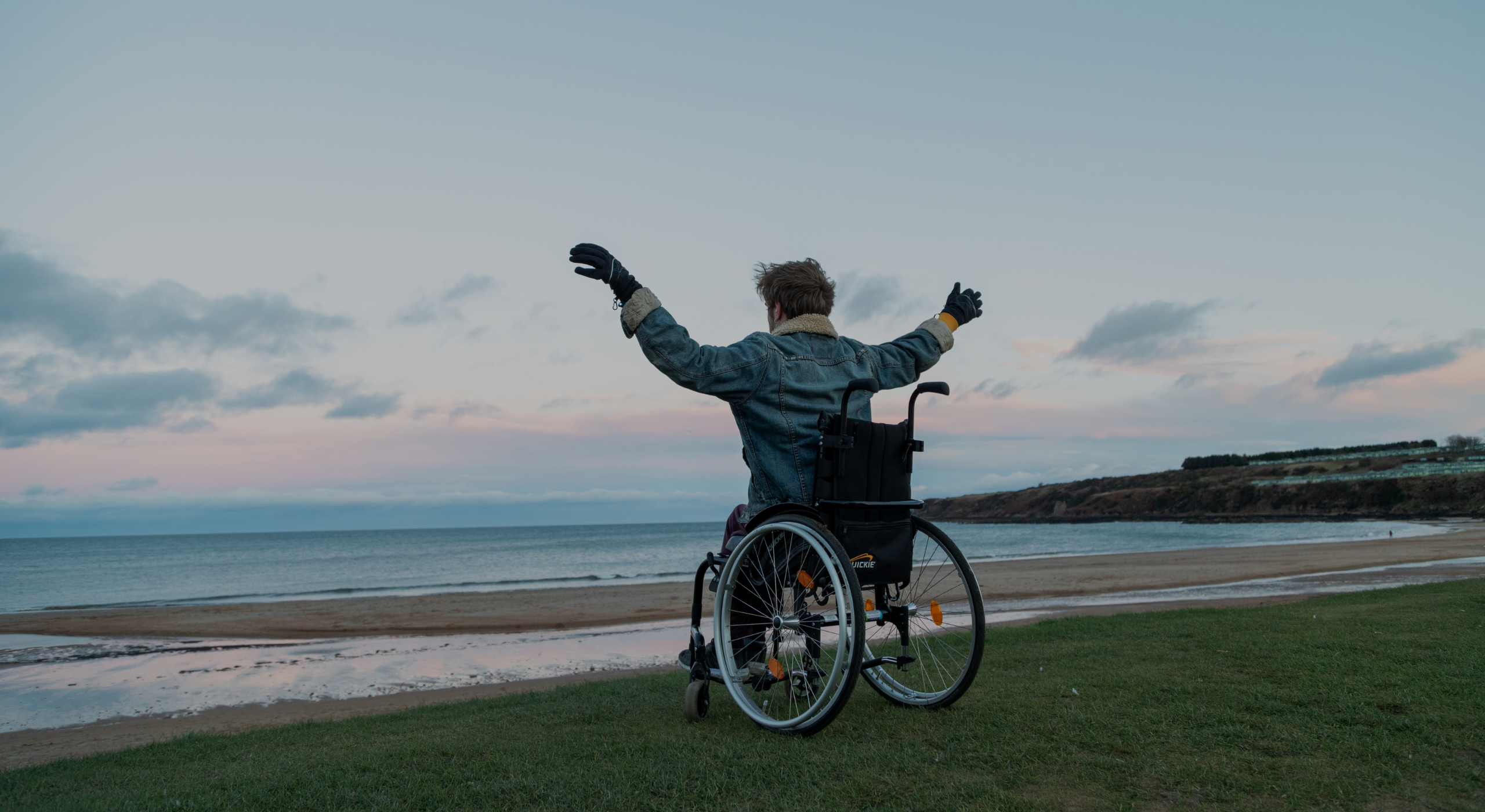 Felix in a wheelchair facing the ocean with arms raised, sitting on grassy ground near a sandy beach under a cloudy sky at sunset.