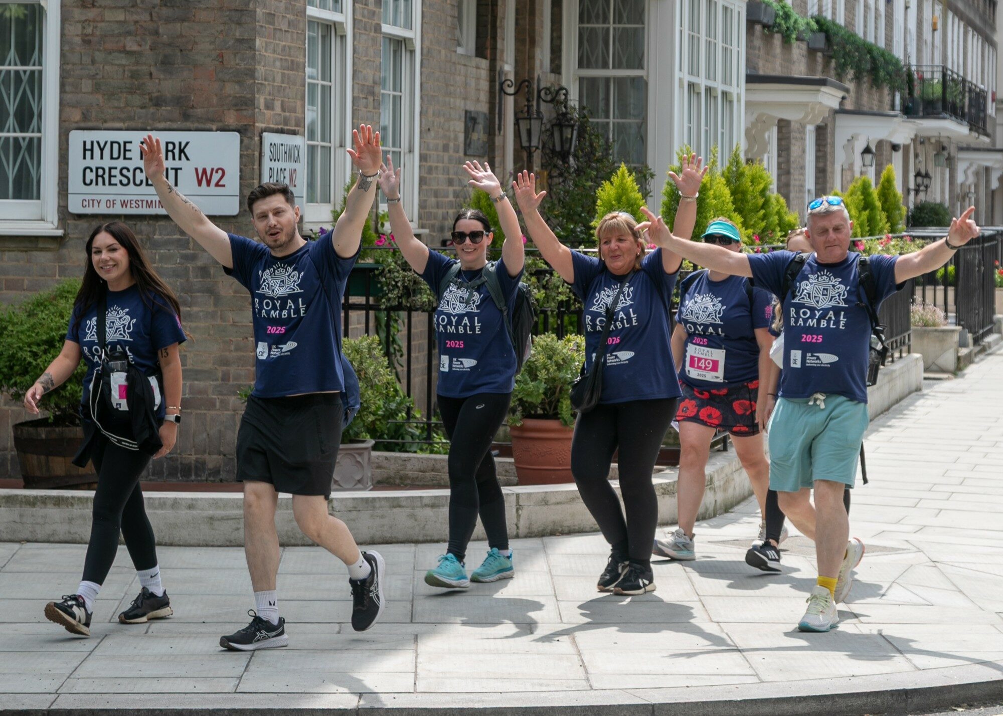 A group of six people walking along a sidewalk in a city, all wearing matching blue t-shirts. They are smiling and raising their arms in a cheerful manner. The background features brick buildings and greenery, with street signs visible.