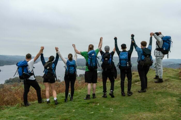 Eight hikers wearing backpacks stand on a grassy hilltop holding hands and raising their arms in celebration facing a scenic view.