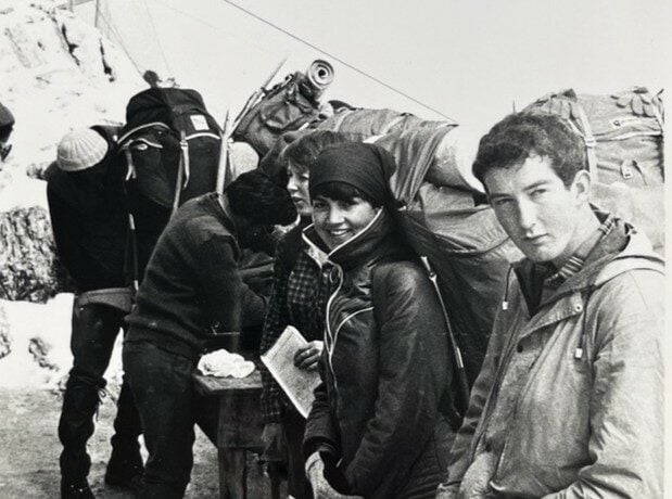 A black‑and‑white 1970s photograph showing a group of people in outdoor clothing preparing for an expedition, surrounded by large rucksacks and equipment in a snowy mountain setting.