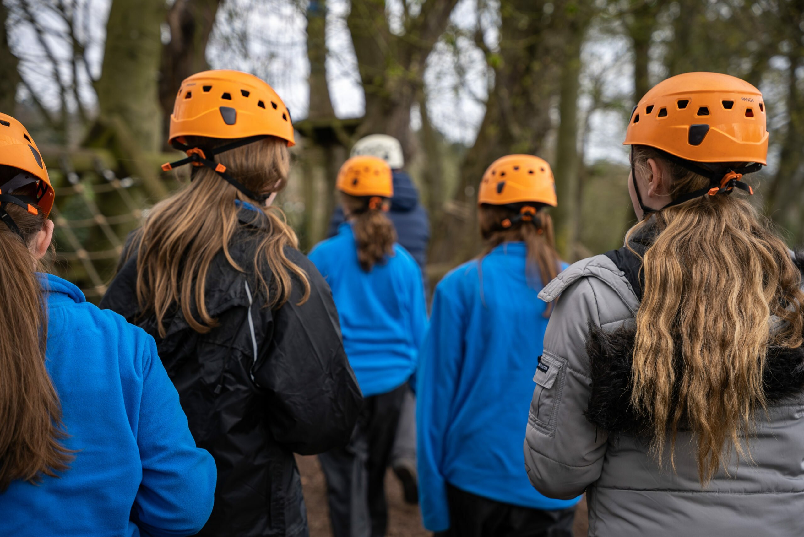 A group of children wearing orange safety helmets and outdoor clothing walk together along a wooded path, surrounded by tall trees and ropes from a climbing or adventure course. The scene is viewed from behind as they move forward in a line.