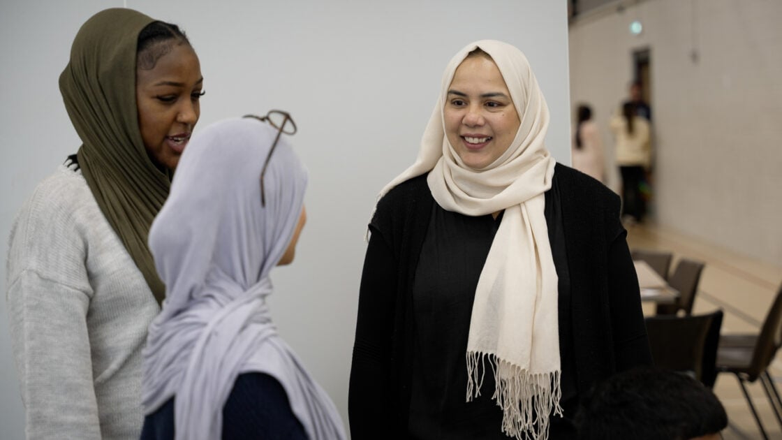 Three women are engaged in conversation indoors. Two women are wearing hijabs in different colors, while the third woman has a light-colored scarf draped over her shoulders. They appear to be smiling and enjoying their discussion, with a blurred background of people and furniture in a communal space.