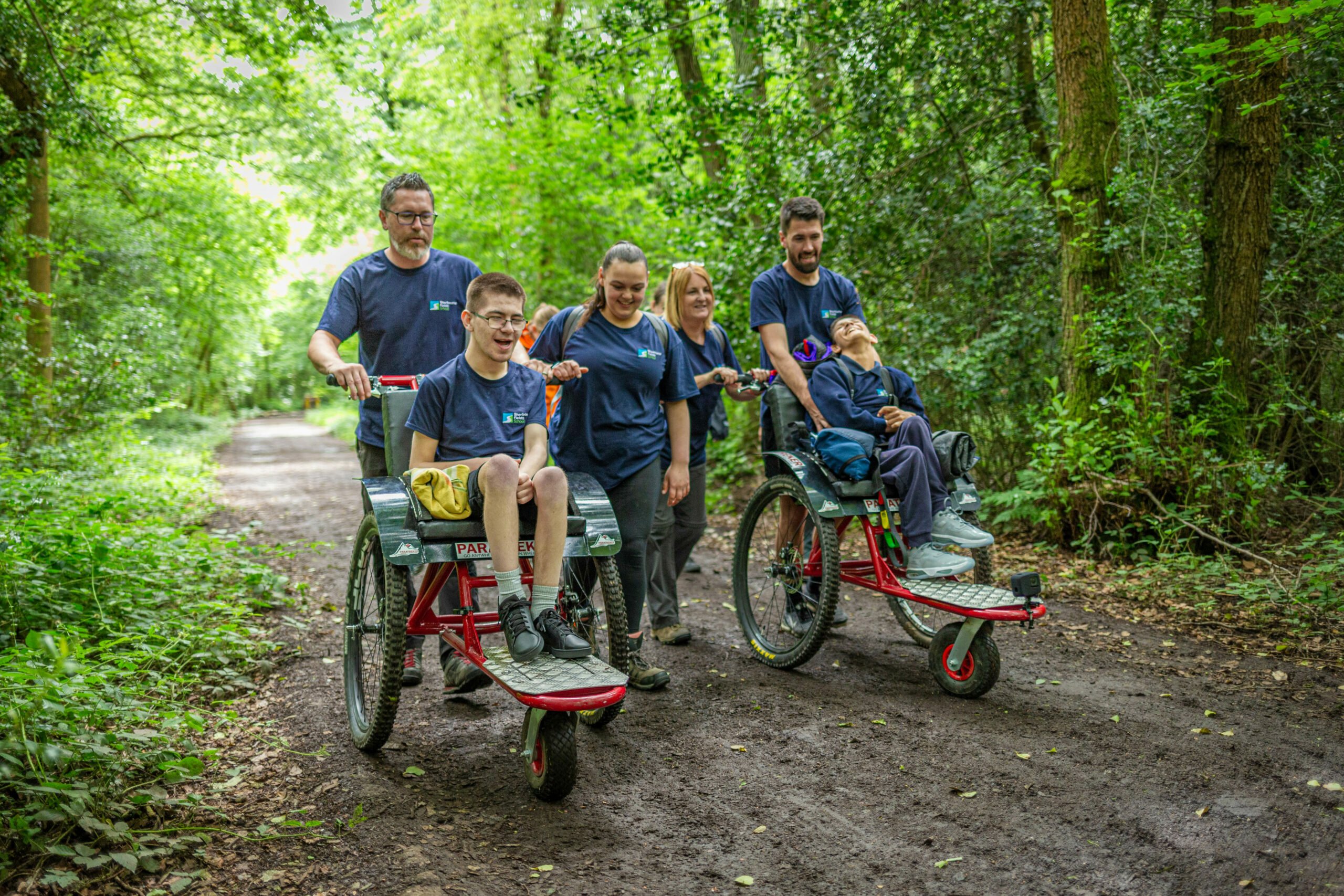 A group of six people is walking along a dirt path in a lush green forest. Two individuals are seated in specialized wheelchairs designed for outdoor use, while four others are accompanying them. Everyone is wearing matching blue t-shirts, and the atmosphere is cheerful and supportive, highlighting a sense of community and inclusivity in nature.