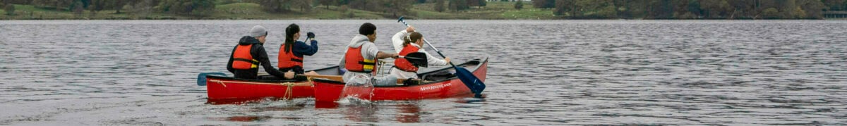 Four people in a canoe paddling across a calm lake, surrounded by lush green trees and rolling hills under a cloudy sky. In the background, a large house is visible on the shore, with mountains rising in the distance.