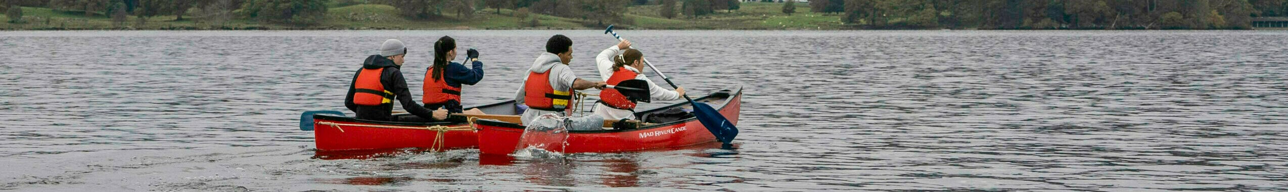 Four people in a canoe paddling across a calm lake, surrounded by lush green trees and rolling hills under a cloudy sky. In the background, a large house is visible on the shore, with mountains rising in the distance.