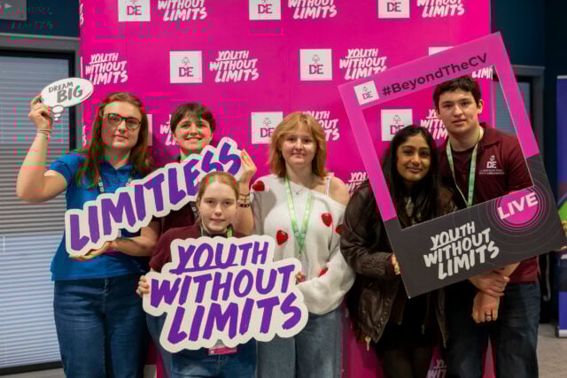 A group of six young people poses in front of a bright pink backdrop with the text 