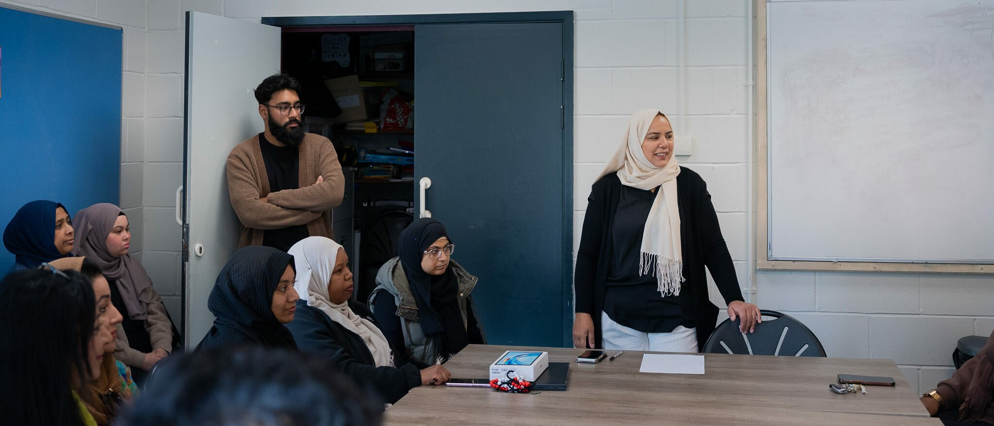 A diverse group of individuals is seated around a table in a classroom setting. A woman wearing a hijab stands at the front, speaking to the group. She appears engaged and confident. In the background, a man with glasses and a sweater leans against the wall, listening attentively. The room has white walls, a whiteboard, and a door leading to another area. The atmosphere is collaborative and focused.