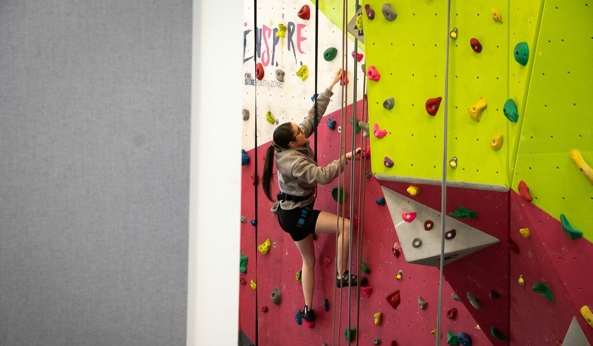 A young person is climbing a colorful indoor rock climbing wall. They are wearing a gray hoodie and shorts, reaching for a hold with one hand while their other hand is positioned on a nearby hold. The wall features various climbing holds in bright colors, including pink, yellow, and green. In the background, part of a gray wall is visible, suggesting the indoor setting.