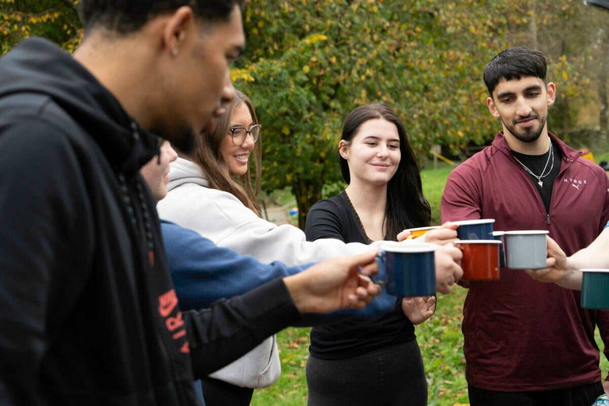 A group of six young adults stands outdoors, smiling and holding up mugs in a toast. The scene is set in a green park with trees in the background. The individuals are dressed casually, and their expressions convey a sense of camaraderie and celebration.
