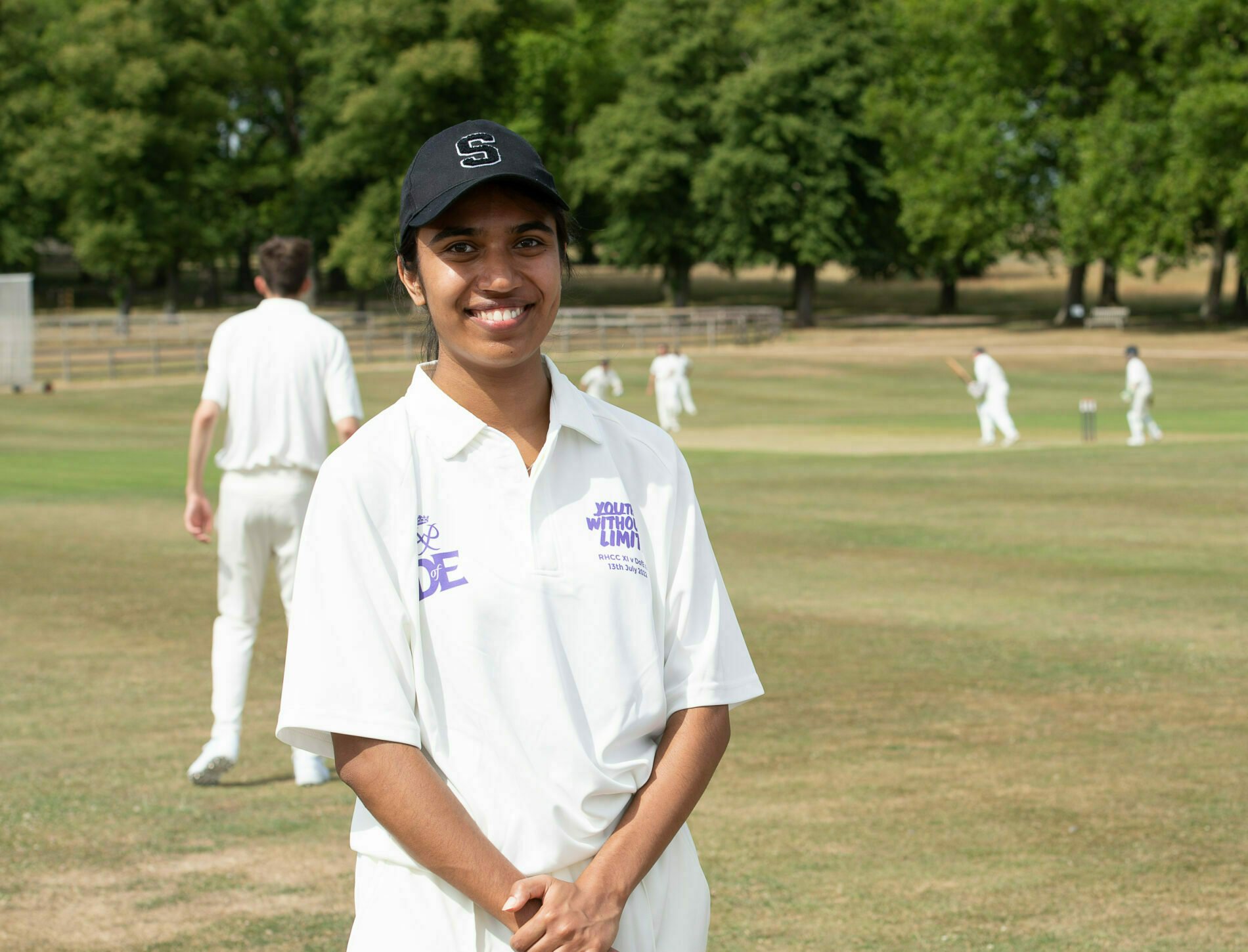 Youth Ambassador, Sonali in a white uniform stands outdoors on a grassy pitch, talking with a man in a white shirt and dark trousers. Other players are visible in the background, along with trees lining the edge of the field.