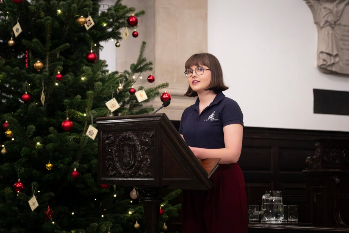 A person stands at a wooden lectern, speaking inside a hall decorated for a festive event. A large Christmas tree adorned with lights and red and gold ornaments stands to the left. The room features dark wood panelling, carved details and a jug of water with glasses on a nearby table.