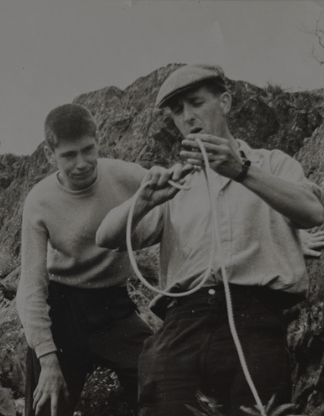 A black‑and‑white photograph of two people outdoors on rocky terrain. One person is demonstrating how to handle or tie a rope, while the other watches closely. Both appear to be engaged in an outdoor skills or climbing activity, with steep rocks visible in the background.