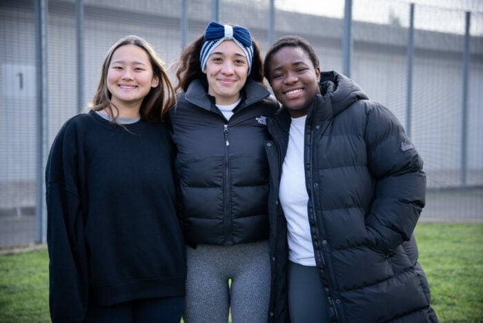 Three young women stand together outdoors, smiling at the camera. They are dressed in casual attire, with one wearing a black sweatshirt, another in a puffy black jacket, and the third in a gray top and leggings. The background features a fenced area, suggesting a sports or recreational setting.