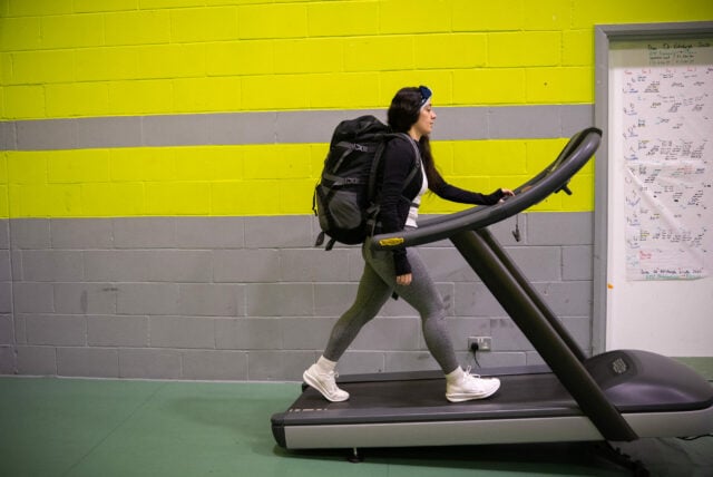 A person with a backpack is walking on a treadmill in a gym. The background features bright green and gray walls, and there are charts or notes on the wall to the right. The individual is dressed in athletic wear, including leggings and a long-sleeve top, and is focused on their workout.