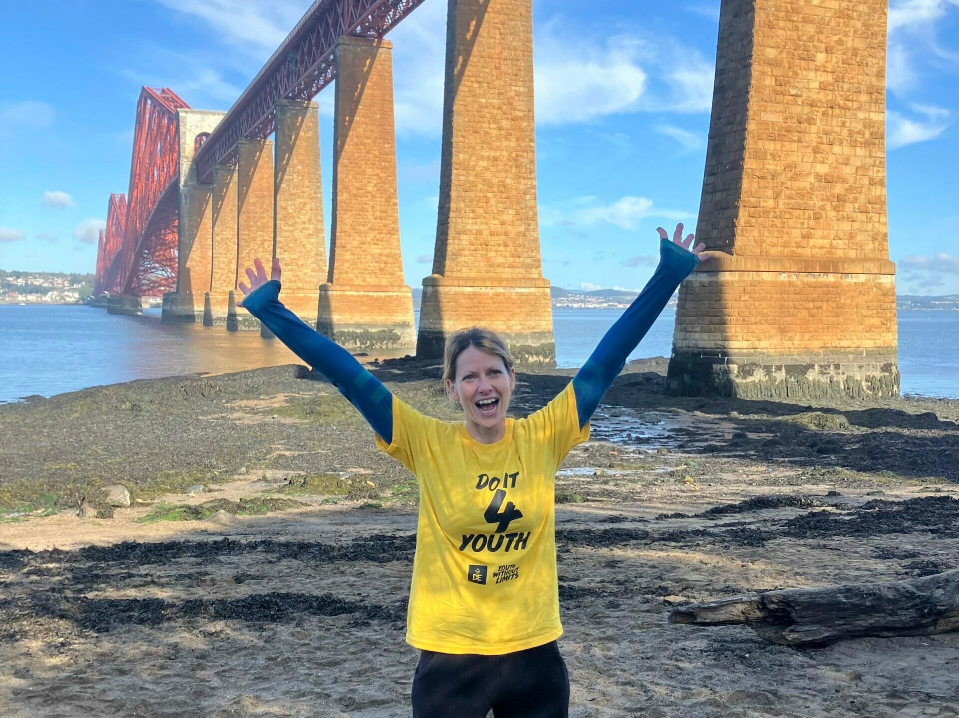 A person in a yellow Do it for youth tshirt holds their hands in the air in front of the Forth Bridge