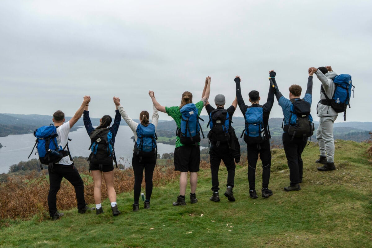A group of eight hikers stands on a grassy hilltop, facing away from the camera. They are holding hands and raising their arms in celebration. Each person is wearing a backpack and dressed in outdoor clothing. In the background, a scenic view of a lake and forested hills is visible under a cloudy sky.