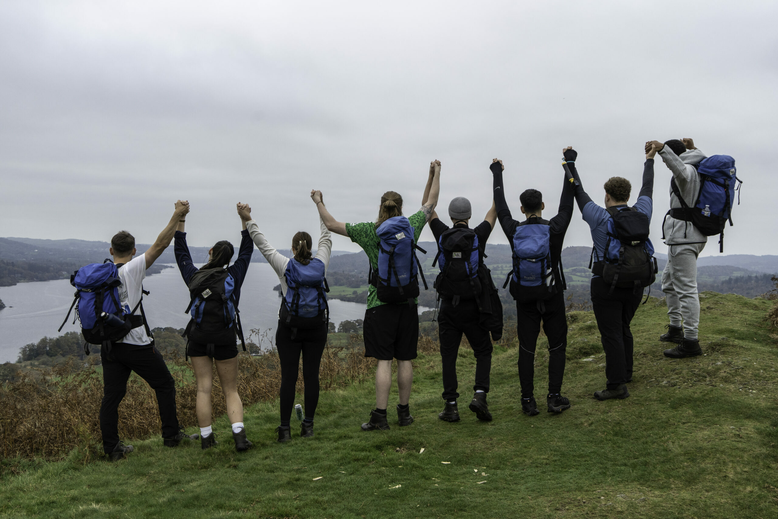 A group of eight hikers stands on a grassy hilltop, facing away from the camera. They are holding hands and raising their arms in celebration. Each person is wearing a backpack and dressed in outdoor clothing. In the background, a scenic view of a lake and forested hills is visible under a cloudy sky.