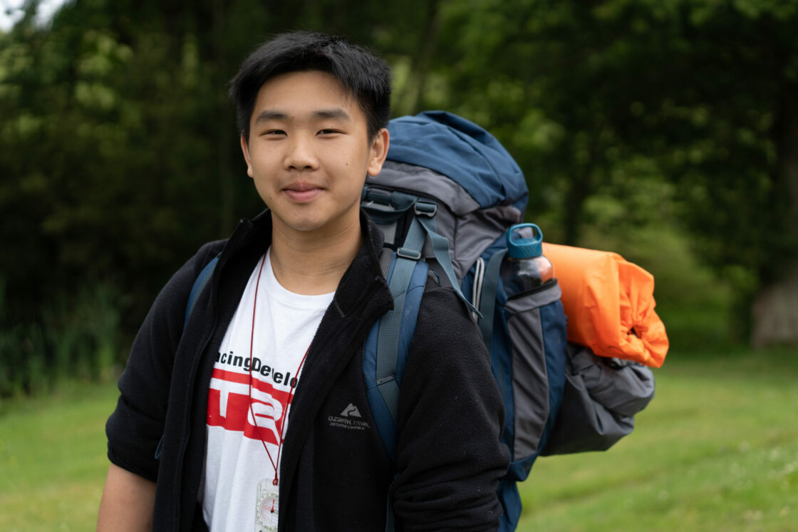 A young man stands outdoors with a backpack and camping gear. He is wearing a white t-shirt and has a slight smile on his face. The background features greenery, suggesting a natural setting.