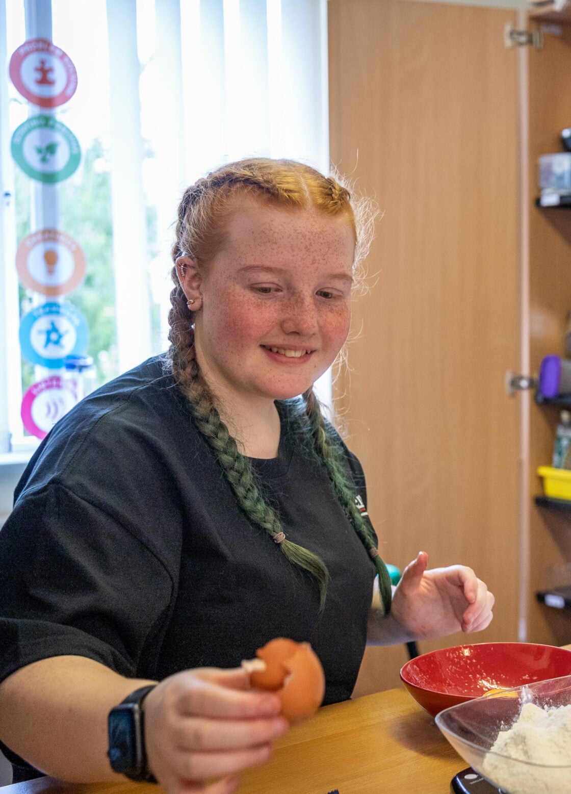Aleea-Mae with long braided hair sits at a table cracking an egg into a bowl. They are wearing a dark T‑shirt and a wristwatch. A red mixing bowl and a clear bowl of flour are on the table. The setting appears to be a bright kitchen or activity room, with colourful circular decorations on the window and shelves in the background.