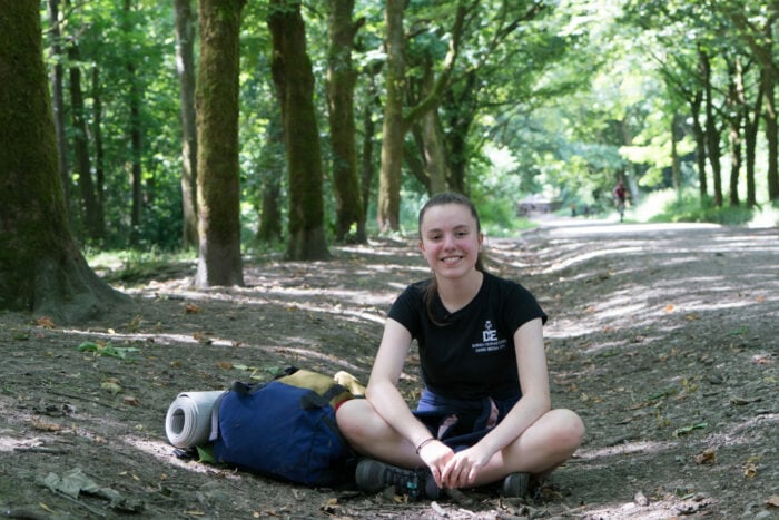 Sarah sits cross legged with a blue rucksack to her right. She is sitting in the middle of a forest with trees either side of her, it is a sunny day.