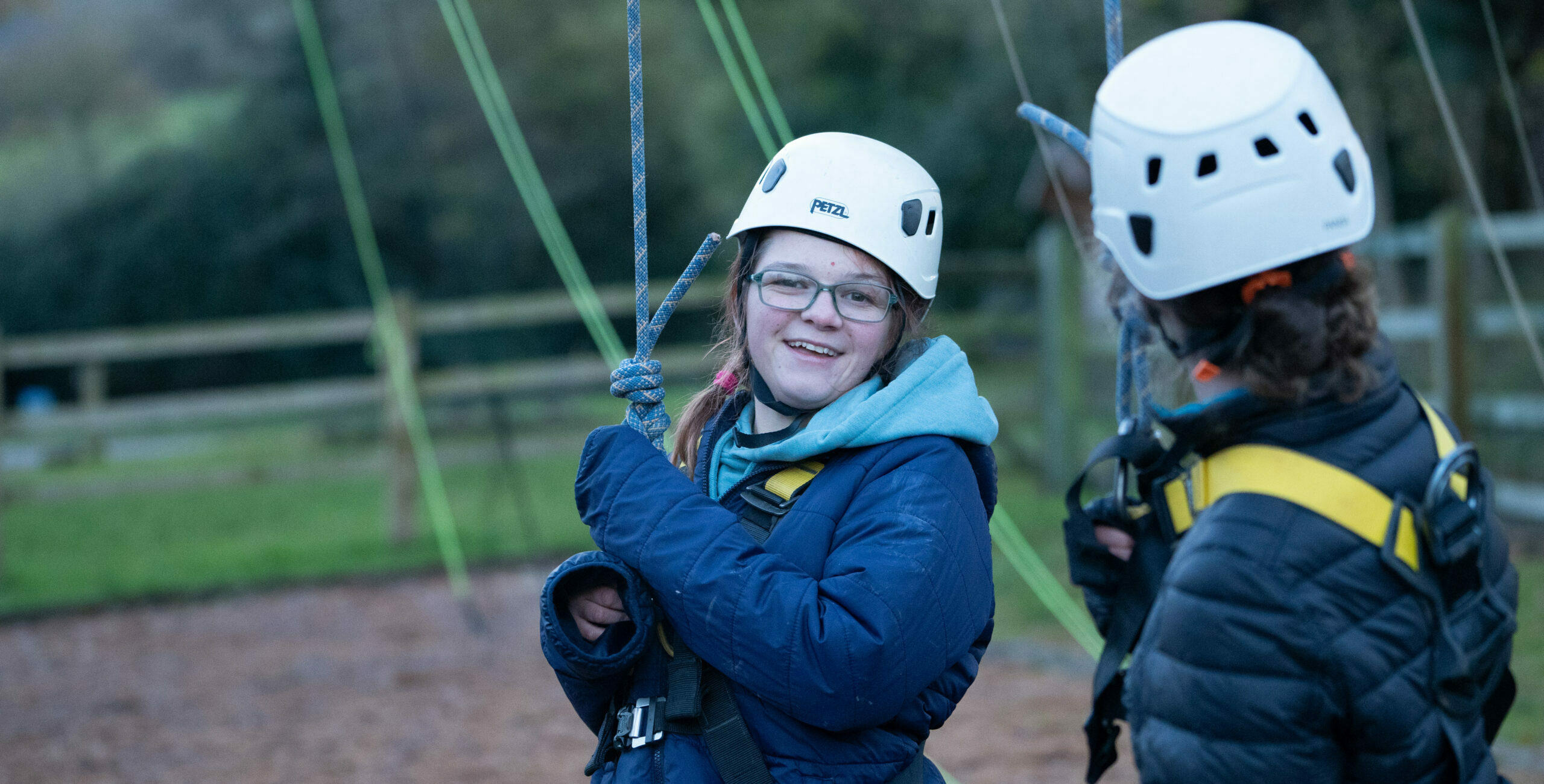 Two young girls are preparing for an outdoor climbing activity. They are wearing helmets and harnesses, with one girl smiling at the camera while the other is looking at her. The background features a grassy area and climbing ropes. The scene is set in a natural environment, suggesting a fun and adventurous atmosphere.