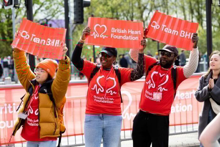 Three people wearing bright red British Heart Foundation T-shirts stand cheering behind a roadside barrier at an outdoor event. They hold up red signs that read “Go Team BHF!” and “British Heart Foundation.” Other spectators stand nearby, and trees and metal barriers line the background.