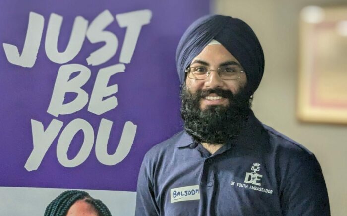 Baljodh wearing a D of E Youth Ambassador polo shirt standing in front of a banner that says 