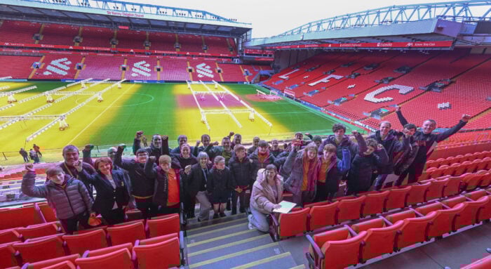 A group of people from D of E and L F C Foundation holding their hands up in celebration at Anfield football stadium.