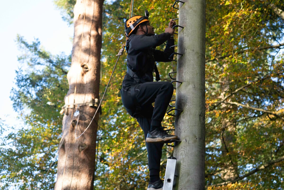 A person wearing a helmet and climbing gear ascends a wooden pole using metal rungs. The background features tall trees with green foliage, indicating an outdoor setting. The individual is focused on their climb, showcasing an adventurous activity.