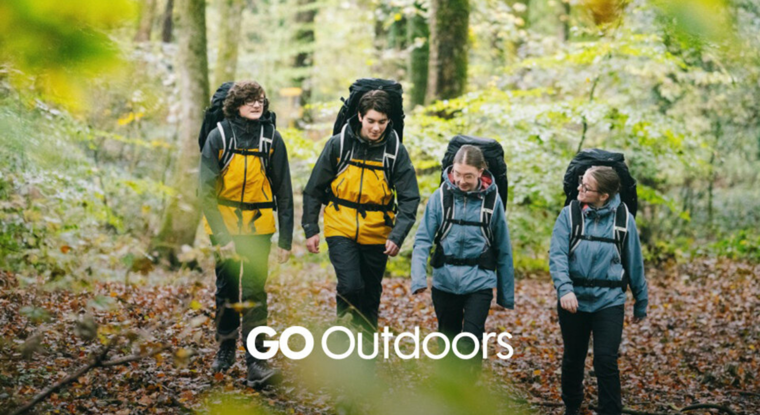 A group of four people wearing outdoor jackets and large backpacks walk together along a leaf‑covered forest trail. The trees around them are green and dense, creating a natural canopy. The individuals appear to be on a hike or expedition. The GO Outdoors logo is displayed at the bottom of the image.