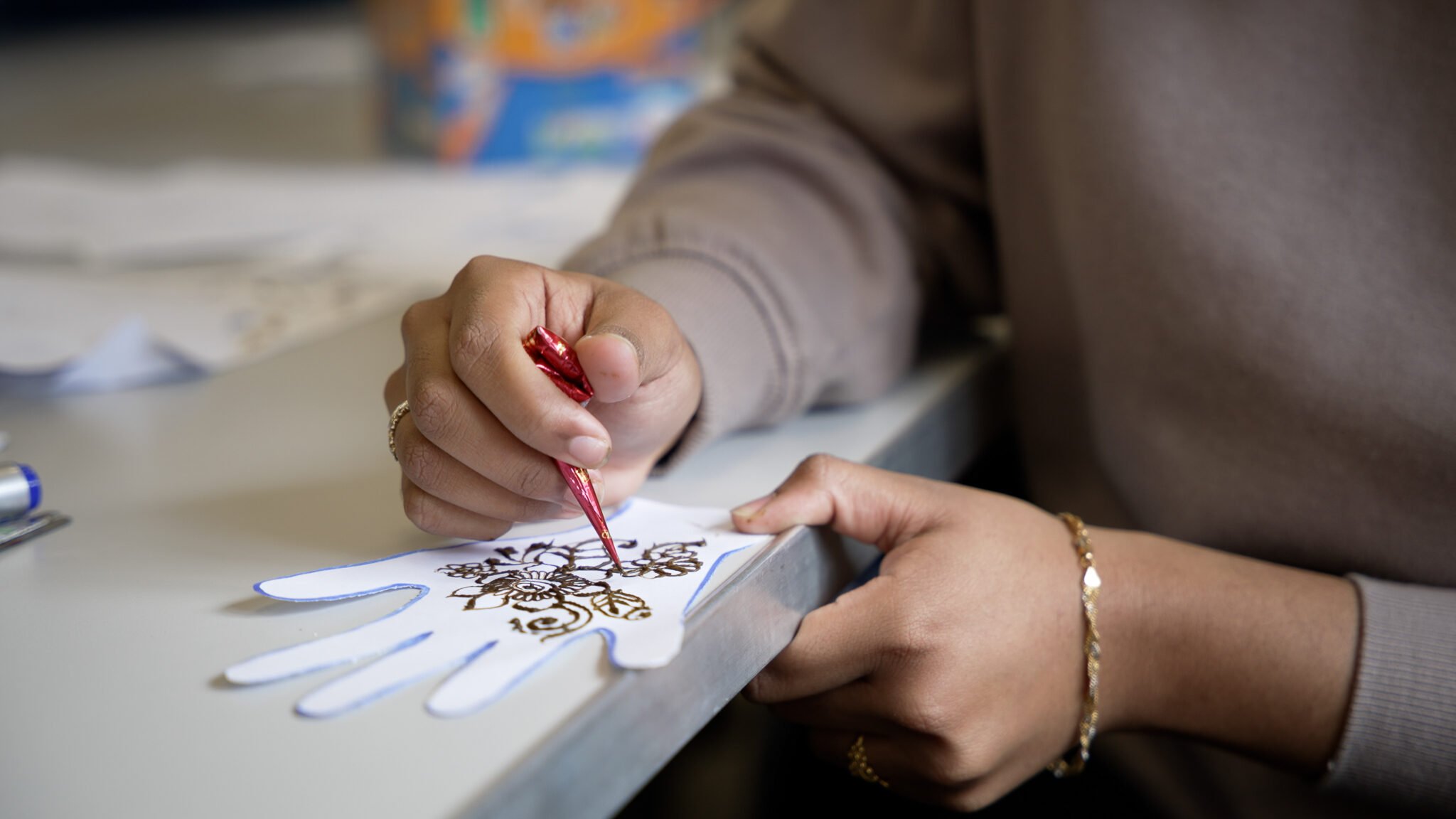 A close-up of a person's hand holding a red paintbrush, carefully painting intricate designs on a white paper cutout shaped like a hand. The background features blurred craft supplies and paper, suggesting a creative workspace.