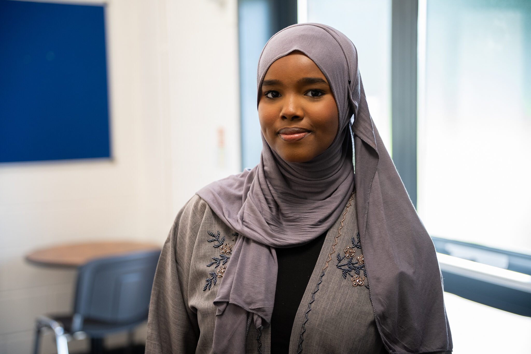 A young woman wearing a light purple hijab and a matching embroidered garment stands in a well-lit room. She has a calm expression and is looking directly at the camera. In the background, there is a blue wall and a table with chairs.
