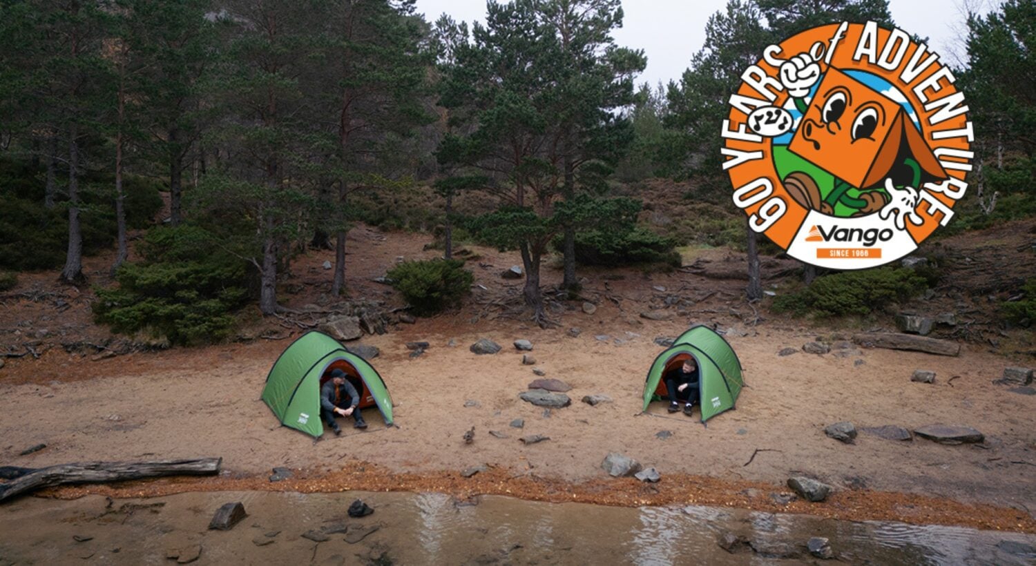 A lakeside campsite with two small green tents pitched on sandy ground near the water’s edge. Each tent has a person seated just inside the entrance. Behind the tents is a dense forest of tall evergreen trees. In the top-right corner, a circular illustrated logo features an orange character wearing a headband with the text “60 Years of Adventure – Vango Since 1966.”