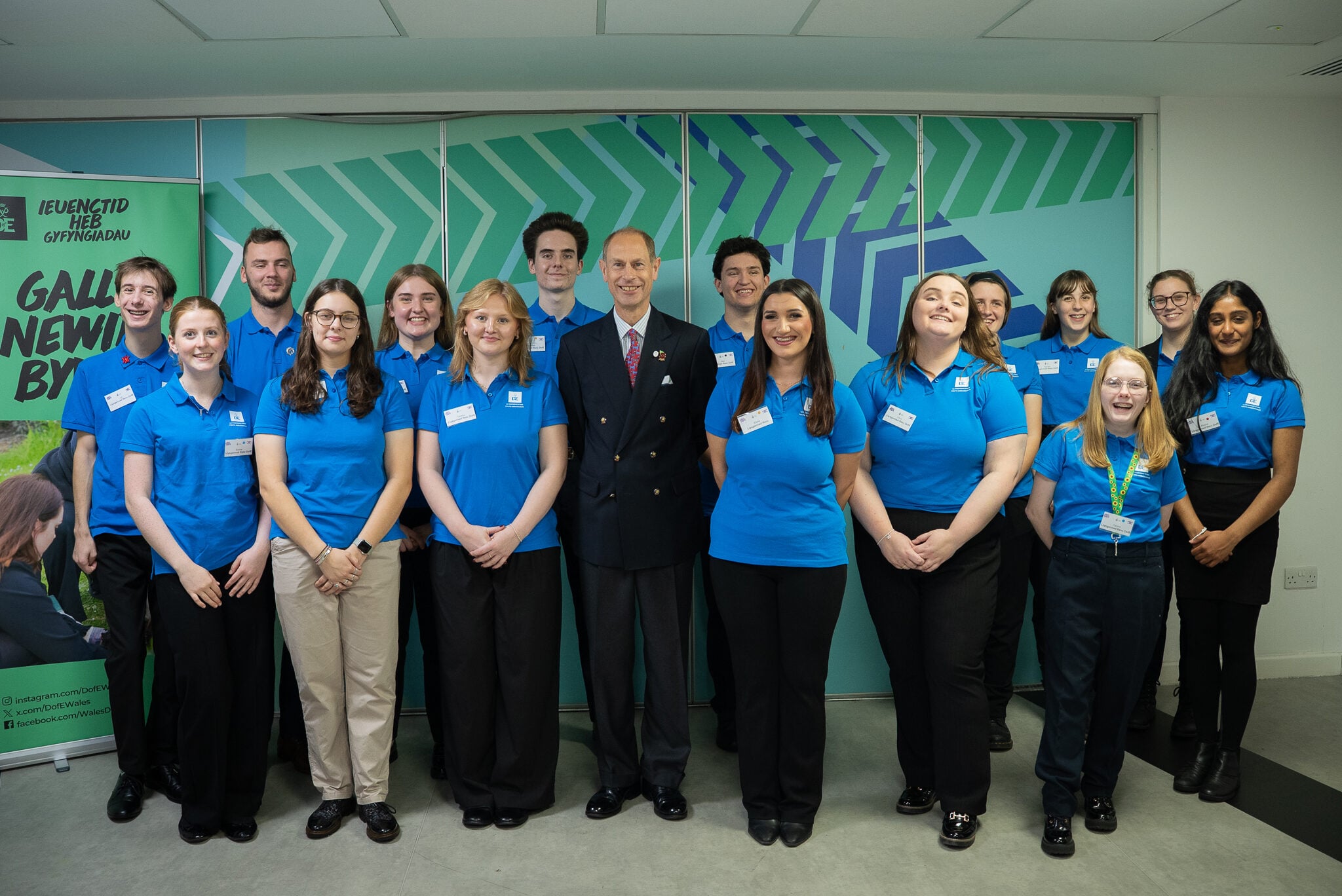A group photo featuring a diverse team of young adults and a HRH The Duke of Edinburgh in a formal uniform, all smiling. They are standing in front of a colorful backdrop with geometric patterns. The young adults are wearing matching blue shirts, while the man in uniform is positioned in the center. The setting appears to be a modern office or event space.