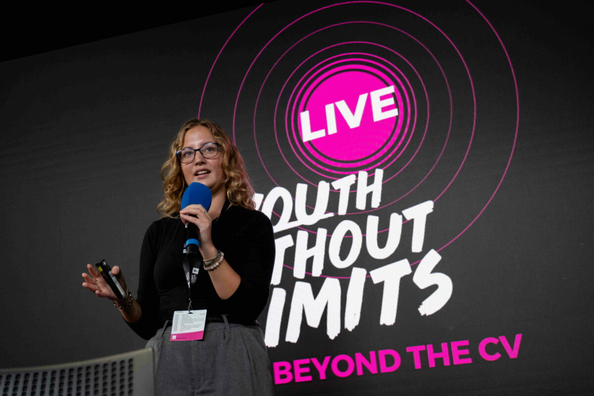 A woman with curly hair stands on stage holding a microphone, speaking to an audience. Behind her is a large screen displaying the text 