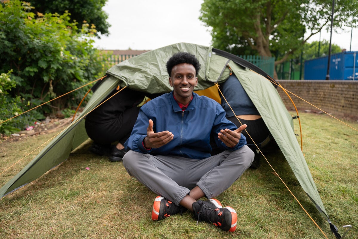 Abdirahman sits on the grass in front of a green tent, smiling and gesturing with his hands. He is wearing a blue jacket and gray pants, with orange and black sneakers. The tent is partially open, revealing two people inside. The background features greenery and a fence.
