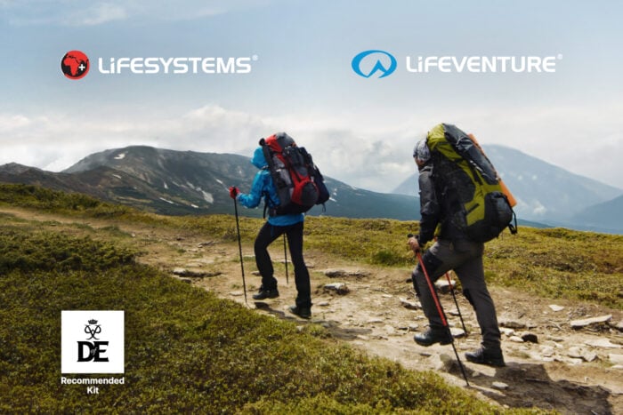 Two hikers wearing large backpacks and using trekking poles walk along a rocky mountain trail surrounded by grassy terrain and distant peaks under a cloudy sky. The Lifesystems and Lifeventure logos appear at the top of the image. A Duke of Edinburgh’s Award ‘Recommended Kit’ badge is shown in the bottom left corner.
