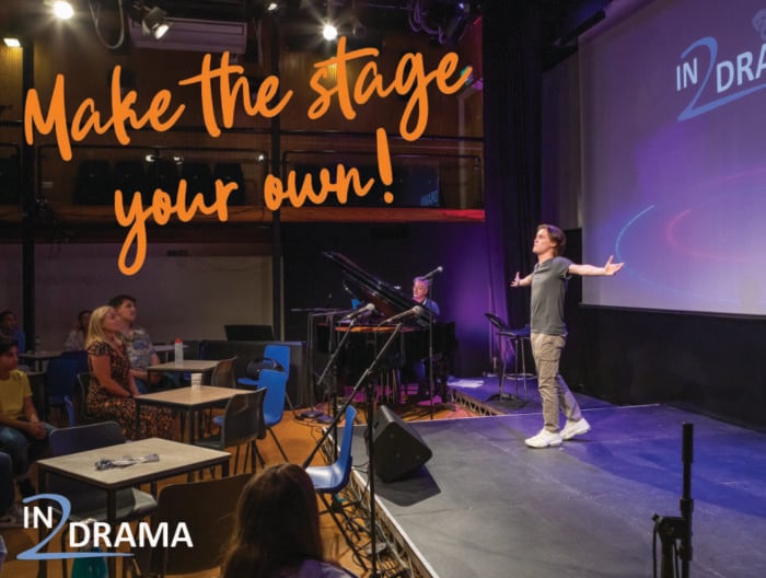 A person stands on a small stage with arms outstretched in a theatre-style room, with an audience seated at tables and a pianist playing nearby. Bold orange text reads “Make the stage your own!” and a large screen displays the “IN2DRAMA” logo. The setting includes stage lighting, musical equipment, and a casual performance atmosphere.