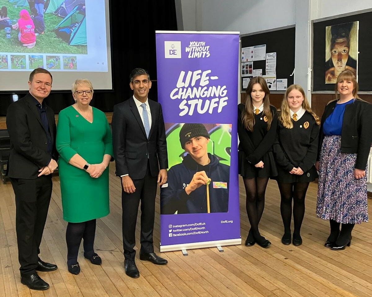 A group of six people stand in a hall beside a purple “Youth Without Limits – Life-Changing Stuff” banner, with school display boards and a projected photo in the background.