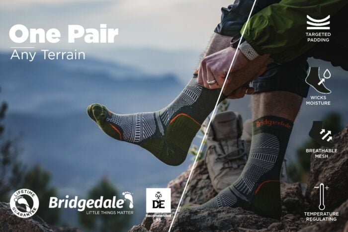 A person sitting on a rocky outdoor surface puts on a pair of Bridgedale hiking socks. The socks are grey with green and orange accents and feature technical patterns. The background shows a soft-focus mountain landscape. Text on the left reads “One Pair – Any Terrain.” Icons on the right highlight features such as targeted padding, moisture‑wicking, breathable mesh, and temperature regulation. Logos along the bottom include Bridgedale, the DofE Recommendation badge, and a Lifetime Guarantee badge.