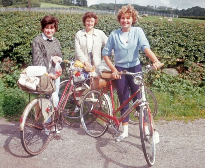 Three women stand beside their bicycles on a gravel path, surrounded by greenery. They are smiling and dressed in casual clothing, with one woman wearing a light blue sweater. Each woman has a bag or basket on her bike, suggesting they are on a leisurely outing. The background features a lush landscape with trees and fields under a partly cloudy sky.