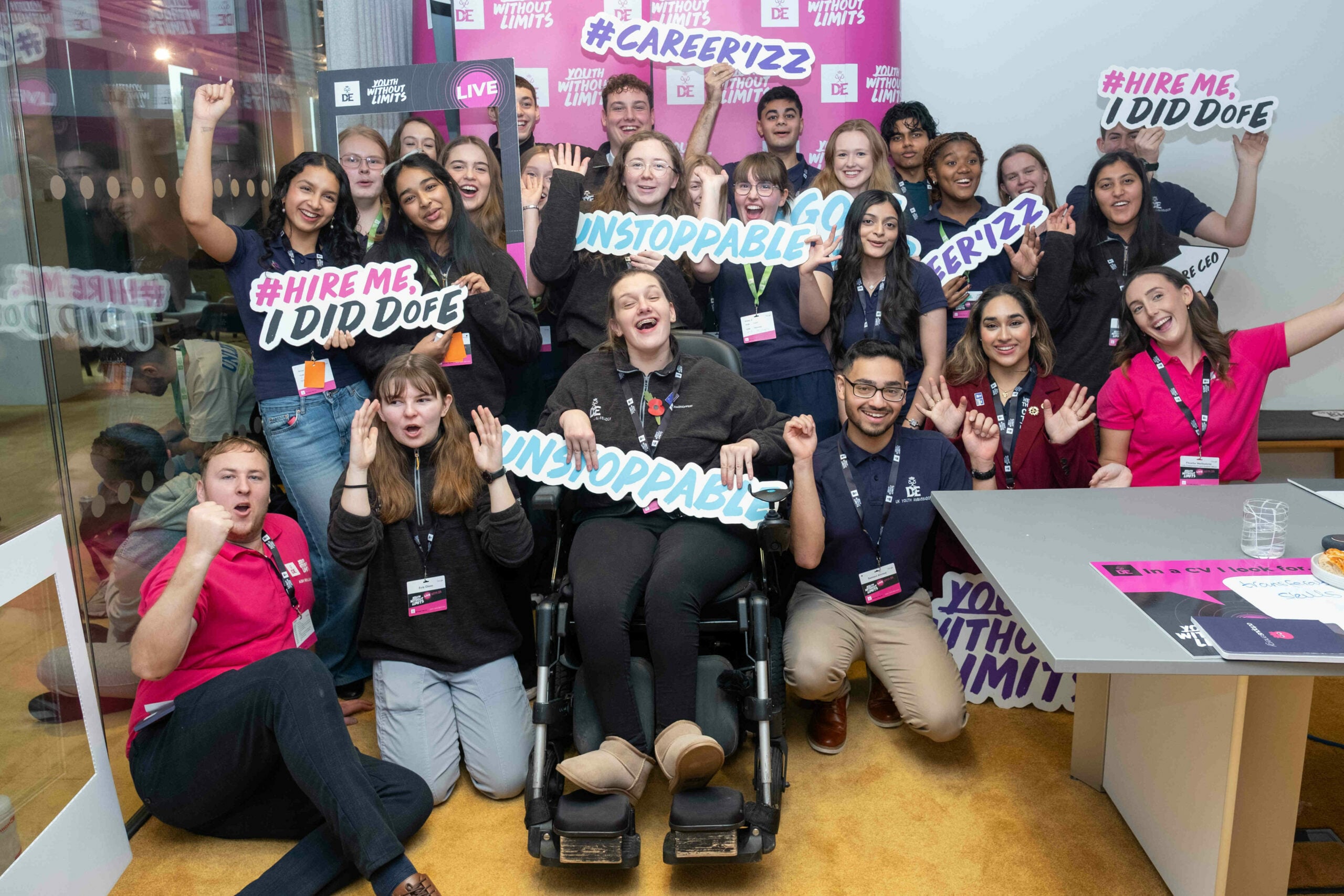 A diverse group of enthusiastic individuals poses together in a brightly lit room, holding up signs with motivational phrases like 