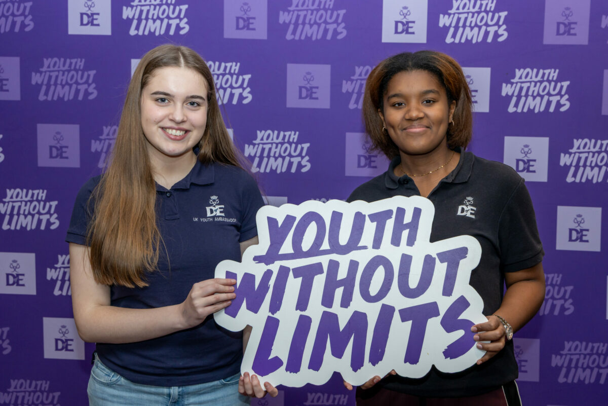 Two young people stand in front of a purple “Youth Without Limits” backdrop, wearing Duke of Edinburgh’s Award polo shirts and holding a sign that reads “Youth Without Limits”.